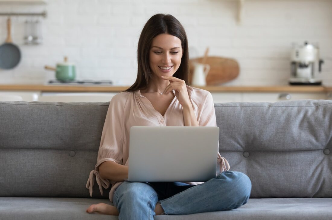 Smiling,Young,Woman,Using,Laptop,,Sitting,On,Couch,At,Home,