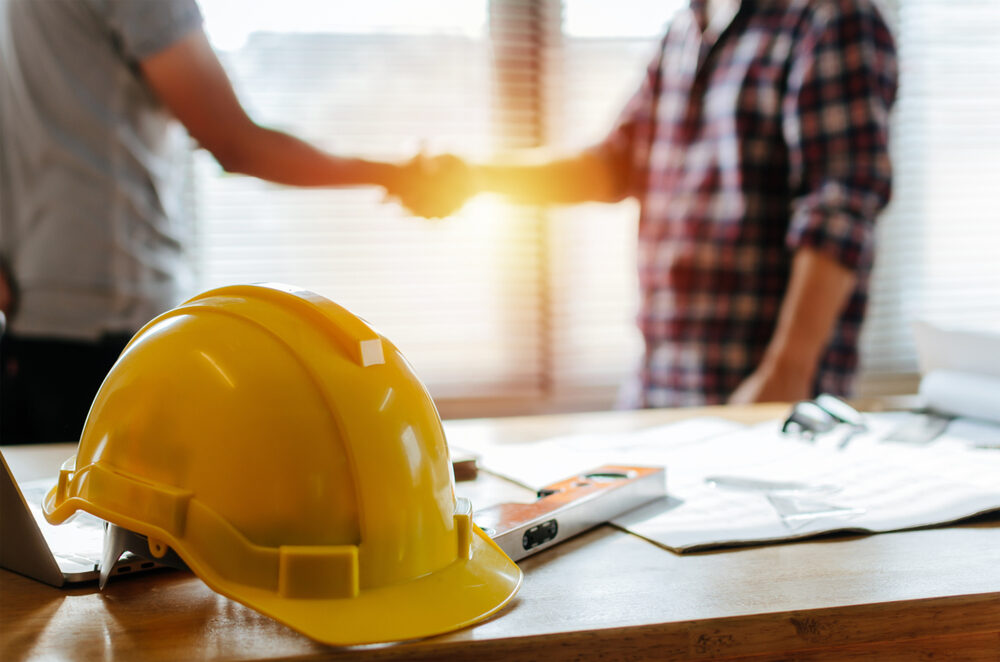 Yellow,Safety,Helmet,On,Workplace,Desk,With,Construction,Worker,Team