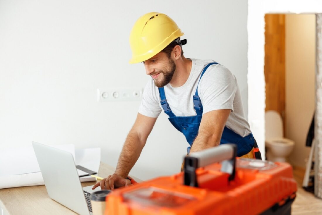 Smiling,Repairman,In,Uniform,And,Hard,Hat,Using,Laptop,,Looking